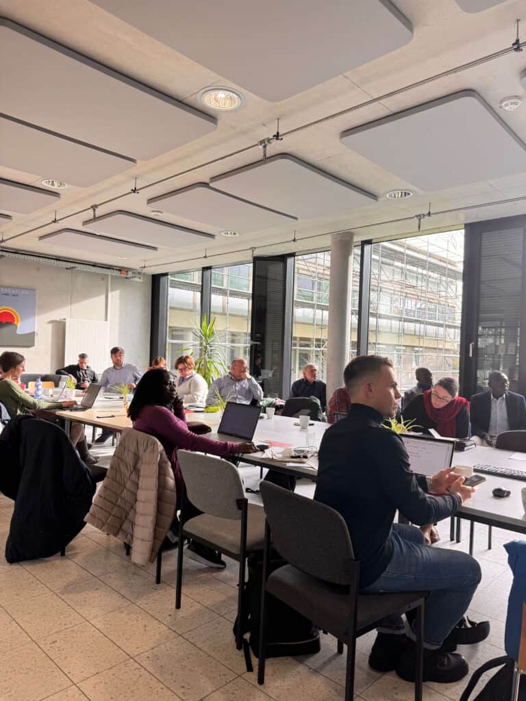 Workshopgäste sitzen in Runde am Tisch und verfolgen einen Vortrag (Vortragende nicht im Bild)